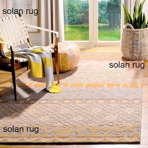Könnte beinhalten: Beige und gelb gemusterter Teppich mit dem Text "solan rug" an mehreren Stellen. Der Teppich befindet sich in einem Raum mit einem Stuhl, einem Pouf und einem Korb mit einer Pflanze. Der Teppich hat ein geometrisches Design mit einem Rand.