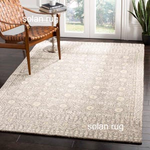 May include: A cream and grey patterned rug, rectangular in shape, with a geometric design. The rug is laid on a dark wooden floor. A leather chair and small table are visible. The rug's pattern includes repeating floral and circular shapes.