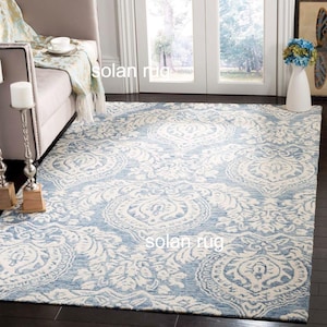 May include: A large, rectangular area rug with a light blue and cream-colored floral pattern. The rug is placed on a dark hardwood floor, with a sofa and decorative items in the background. The rug's design features a repeating medallion motif.