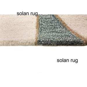 May include: Close-up of a textured rug with a geometric design. The rug features a cream-colored section, a section of teal, and a brown border. The words "solan rug" are printed on the rug.