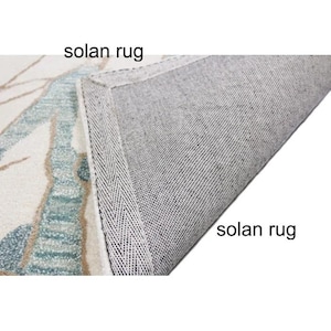 May include: A section of a rug with a floral design in shades of blue, beige, and white. The underside of the rug is gray, and the text "solan rug" is visible in two places.