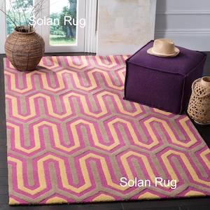 Könnte beinhalten: Ein Teppich mit geometrischem Muster in Rosa, Beige und Grau. Ein violetter quadratischer Ottoman mit einem Strohhut liegt darauf. Eine geflochtene Vase und eine dekorative Vase sind ebenfalls im Bild. Der Text "Solan Rug" ist sichtbar.