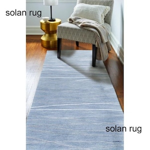 May include: A light blue Solan rug with white linear patterns. The rug is rectangular and placed on a hardwood floor. The text "solan rug" is visible in the image. A patterned chair, a side table, and a lamp are in the background.