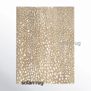 May include: A rectangular rug with a tan background and a pattern of white, irregular spots. The rug has a speckled design, resembling animal hide. The words "solan rug" are printed in black at the bottom and right side of the rug.