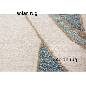 May include: Close-up of a cream-colored rug with a textured surface. The rug features a design with teal and brown geometric shapes. The text "solan rug" is printed on the rug.