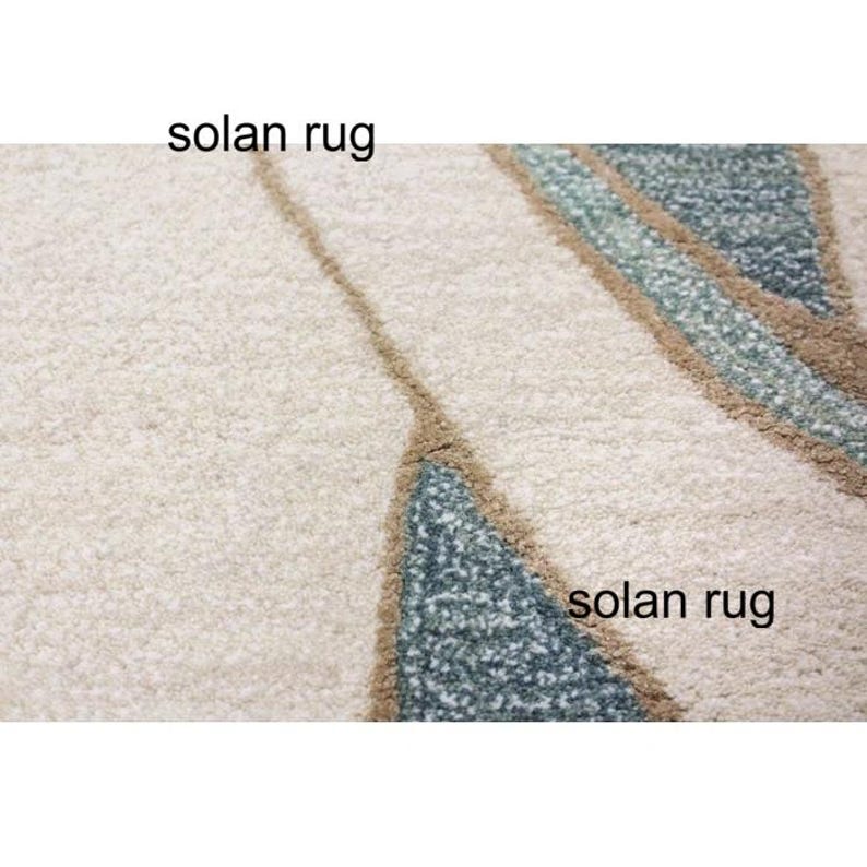 May include: Close-up of a cream-colored rug with a textured surface. The rug features a design with teal and brown geometric shapes. The text "solan rug" is printed on the rug.