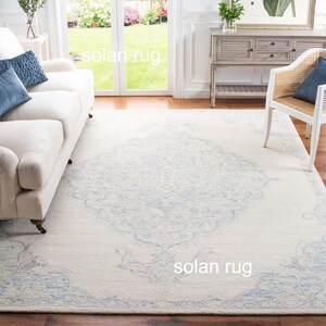 May include: A large, rectangular rug with a cream background and a detailed, light blue floral pattern. The rug is in a living room, with a white sofa and a wooden chair visible. The rug's dimensions are in centimetres.