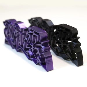 Könnte beinhalten: Zwei 3D-gedruckte Dekorationsgegenstände. Einer ist leuchtend lila, der andere schwarz. Beide haben ein komplexes, geometrisches Design. Die Objekte sind auf einer weißen Oberfläche platziert, wobei sich das lila Objekt im Vordergrund befindet.