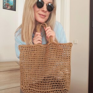 May include: A tan-colored, rectangular tote bag crafted from a coarse, open-weave material. The bag features a grid-like pattern throughout its body and includes two integrated handles at the top, allowing for easy carrying of the open-top accessory.