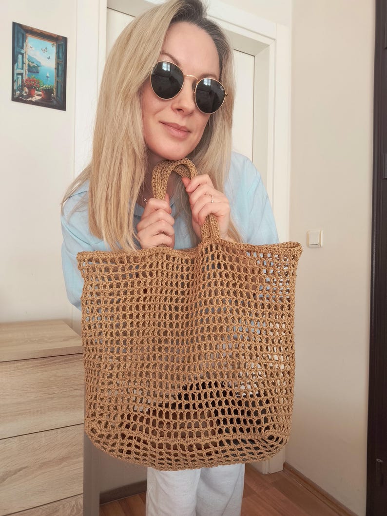 May include: A tan-colored, rectangular tote bag crafted from a coarse, open-weave material. The bag features a grid-like pattern throughout its body and includes two integrated handles at the top, allowing for easy carrying of the open-top accessory.