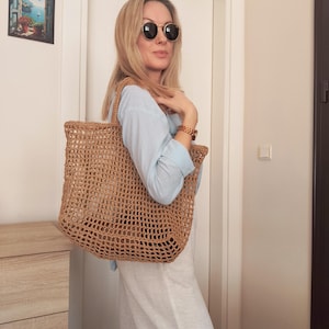 May include: A large, tan, open-weave crochet tote bag with two shoulder straps. The bag features a grid-like pattern constructed from natural-colored fiber, providing a spacious interior compartment for carrying items while maintaining a lightweight structure.