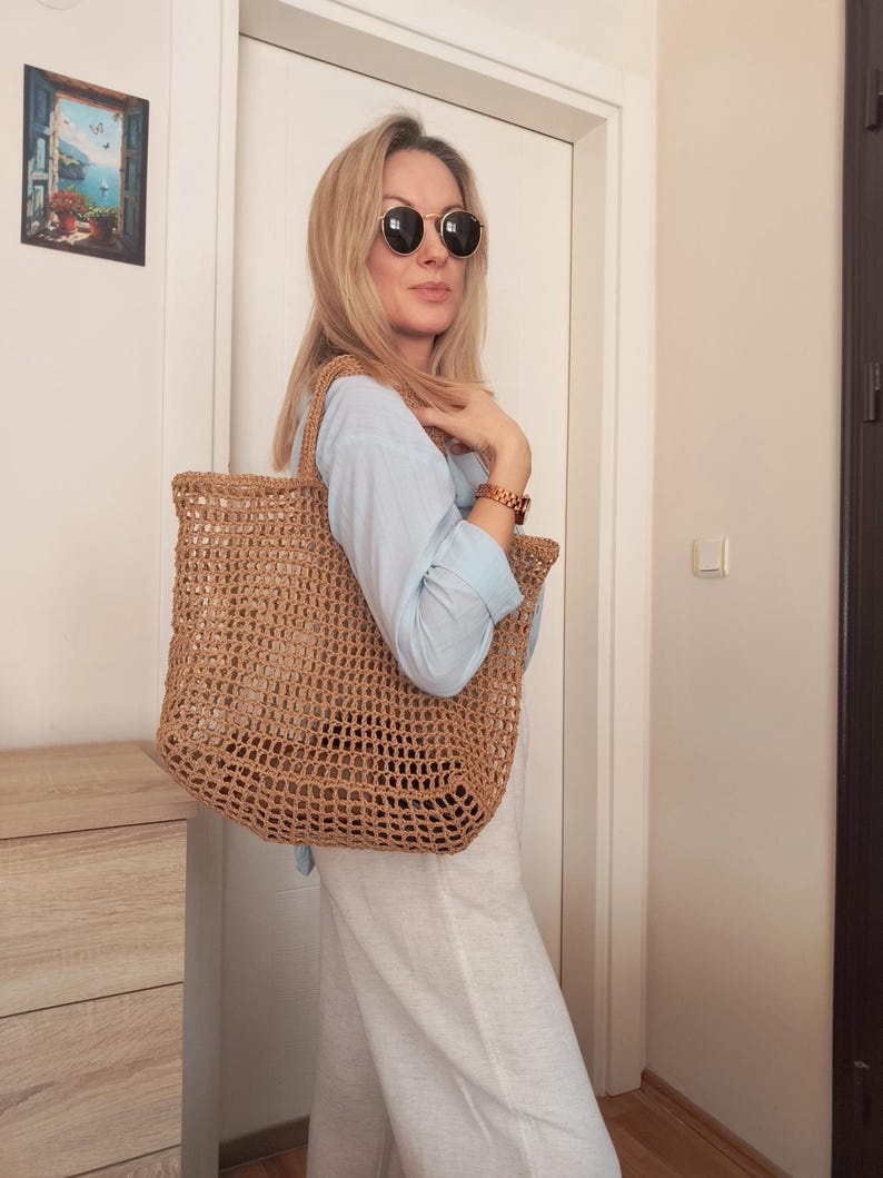 May include: A large, tan, open-weave crochet tote bag with two shoulder straps. The bag features a grid-like pattern constructed from natural-colored fiber, providing a spacious interior compartment for carrying items while maintaining a lightweight structure.