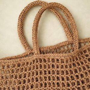 May include: A tan-colored tote bag crafted with a crochet mesh construction. The bag features two sturdy, rounded handles attached to the top edge, with a consistent open-weave grid pattern throughout the body of the bag, showcasing the fiber texture.