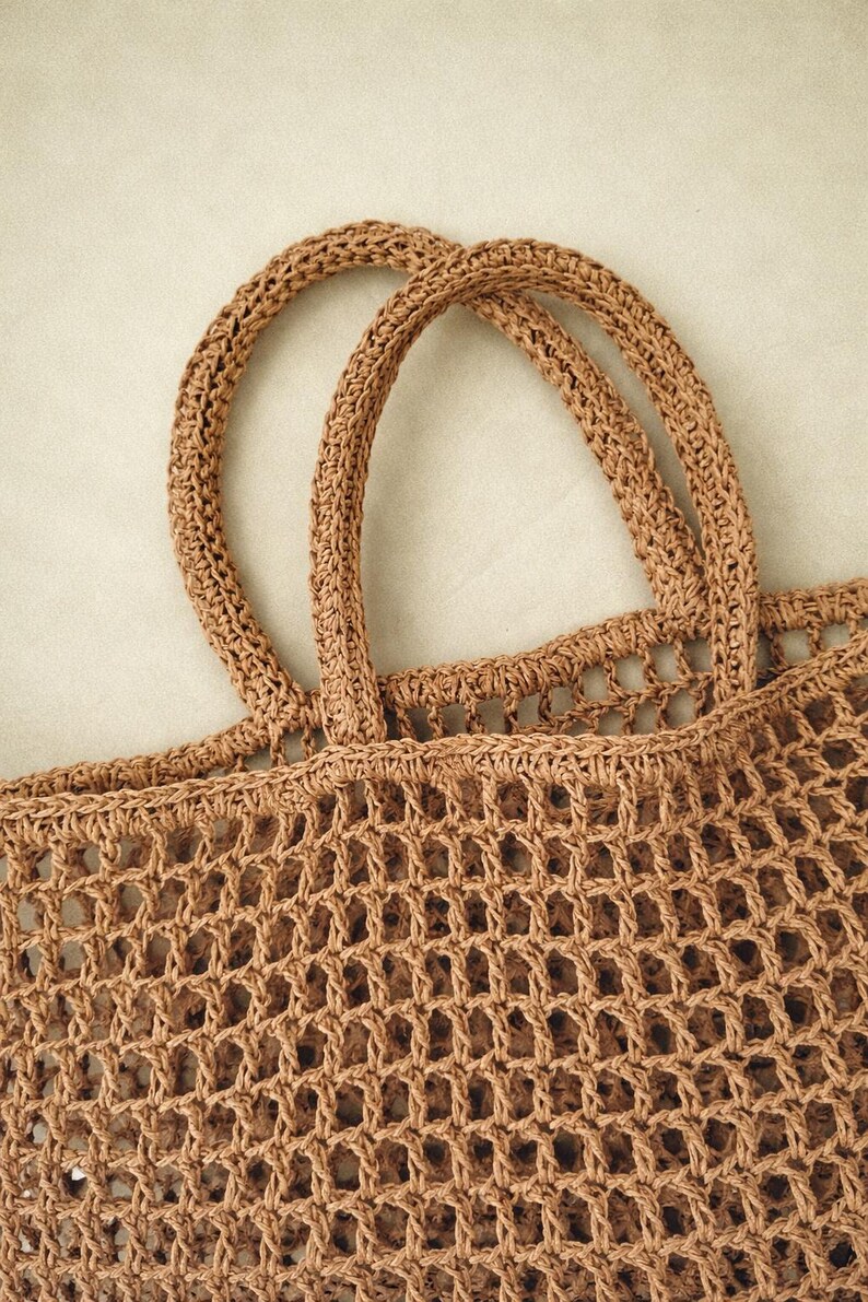 May include: A tan-colored tote bag crafted with a crochet mesh construction. The bag features two sturdy, rounded handles attached to the top edge, with a consistent open-weave grid pattern throughout the body of the bag, showcasing the fiber texture.