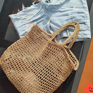 May include: A light tan, open-weave crochet tote bag with two matching handles, positioned next to a pair of light blue denim shorts featuring a distressed, frayed hemline and visible white pocket lining through the ripped fabric sections.