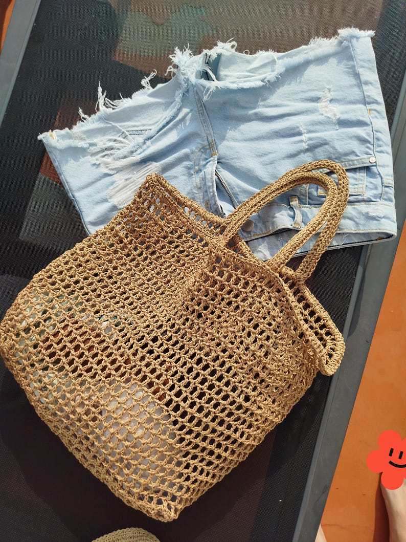 May include: A light tan, open-weave crochet tote bag with two matching handles, positioned next to a pair of light blue denim shorts featuring a distressed, frayed hemline and visible white pocket lining through the ripped fabric sections.