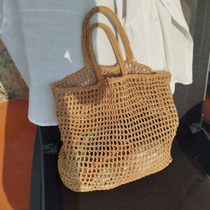 May include: A tan crochet tote bag featuring an open-weave grid pattern, two integrated top handles, and a rectangular base. The bag is constructed from a textured, straw-like fiber and displays a consistent light brown color throughout the design.