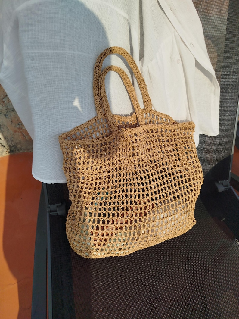 May include: A tan crochet tote bag featuring an open-weave grid pattern, two integrated top handles, and a rectangular base. The bag is constructed from a textured, straw-like fiber and displays a consistent light brown color throughout the design.