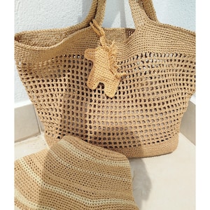 Op de afbeelding: Een gehaakte tote bag en bijpassende zonnehoed in een natuurlijke beige kleur. De tas heeft een groot, open geweven ontwerp en stevige handvatten. Een kleine, decoratieve paardvormige bedel hangt aan de tas. De hoed heeft horizontale strepen.