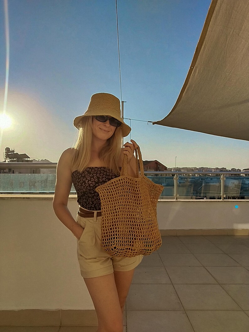 May include: A tan crochet tote bag with an open-weave mesh design and two top handles. The bag is constructed from a textured, natural-colored fiber, featuring a spacious, flexible body with a consistent grid pattern throughout the entire structure.