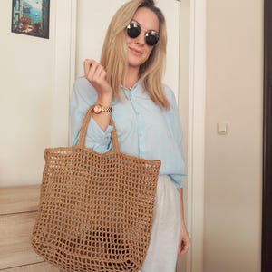 May include: A large, rectangular, tan-colored tote bag crafted from a coarse, open-weave natural fiber material. The bag features two sturdy, matching woven handles at the top and a spacious, unlined interior compartment with a visible grid pattern.