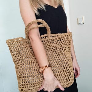 May include: A large, tan-colored tote bag crafted from a woven, open-grid material. The bag features two sturdy, matching woven handles at the top and a rectangular base, showcasing a consistent, textured, and airy construction throughout.