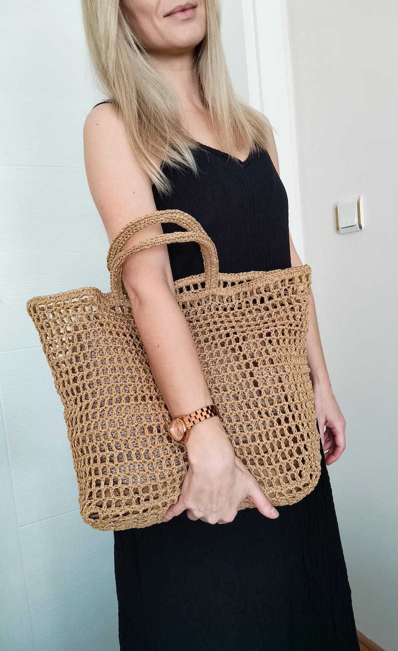 May include: A large, tan-colored tote bag crafted from a woven, open-grid material. The bag features two sturdy, matching woven handles at the top and a rectangular base, showcasing a consistent, textured, and airy construction throughout.
