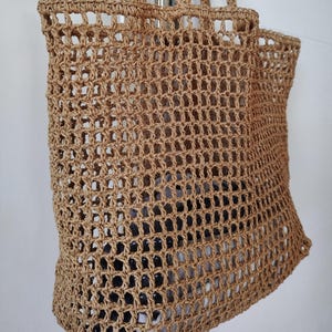 May include: A tan-colored tote bag crafted from a woven, open-grid material. The bag features two sturdy handles at the top and a rectangular, structured base. The open-weave design reveals a glimpse of a dark, patterned item stored inside the bag.