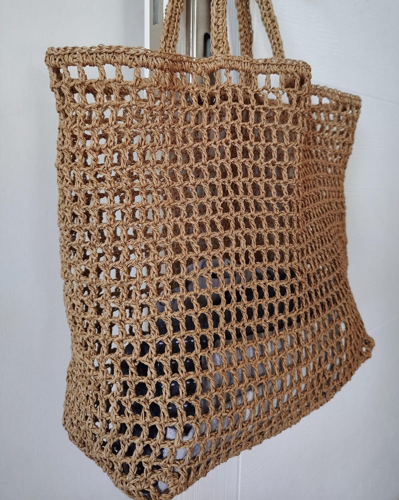 May include: A tan-colored tote bag crafted from a woven, open-grid material. The bag features two sturdy handles at the top and a rectangular, structured base. The open-weave design reveals a glimpse of a dark, patterned item stored inside the bag.