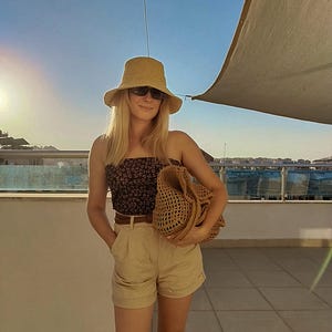 May include: A tan straw bucket hat, dark sunglasses, a black tube top with a small white floral pattern, a brown belt, tan high-waisted shorts with a cuffed hem, and a large tan woven straw tote bag with an open-knit texture and wide handles.