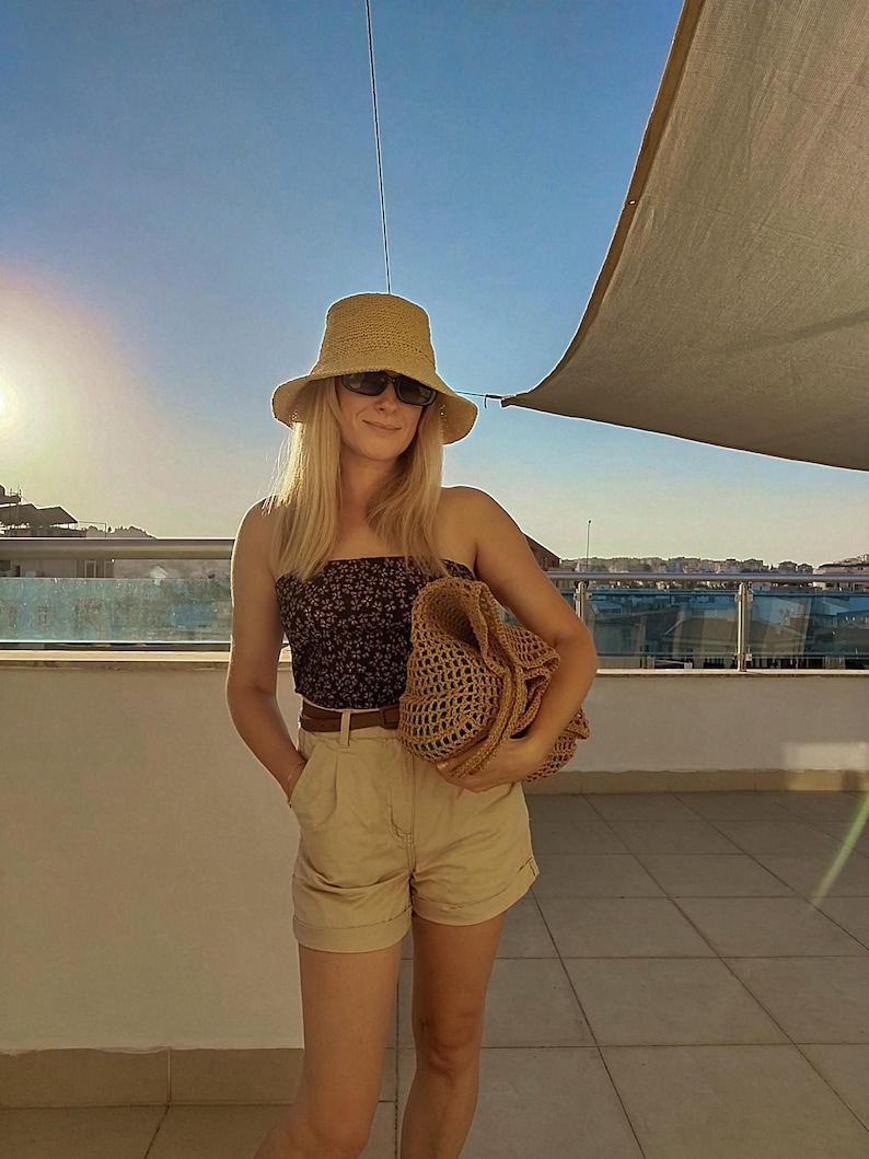 May include: A tan straw bucket hat, dark sunglasses, a black tube top with a small white floral pattern, a brown belt, tan high-waisted shorts with a cuffed hem, and a large tan woven straw tote bag with an open-knit texture and wide handles.