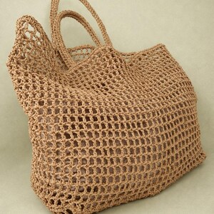 May include: A tan crochet tote bag featuring an open-weave grid pattern and two matching top handles. The bag has a structured rectangular base and a soft, flexible body, showcasing a consistent light brown color throughout the entire construction.