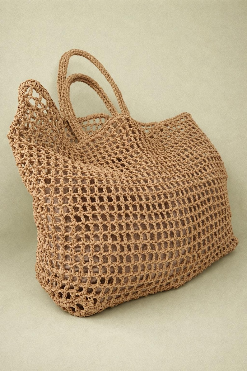 May include: A tan crochet tote bag featuring an open-weave grid pattern and two matching top handles. The bag has a structured rectangular base and a soft, flexible body, showcasing a consistent light brown color throughout the entire construction.