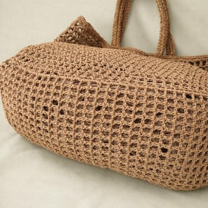 May include: A light brown crochet tote bag featuring an open grid pattern and two sturdy handles. The bag has a rounded, structured base and a visible inner lining that provides a solid appearance through the woven exterior mesh construction.