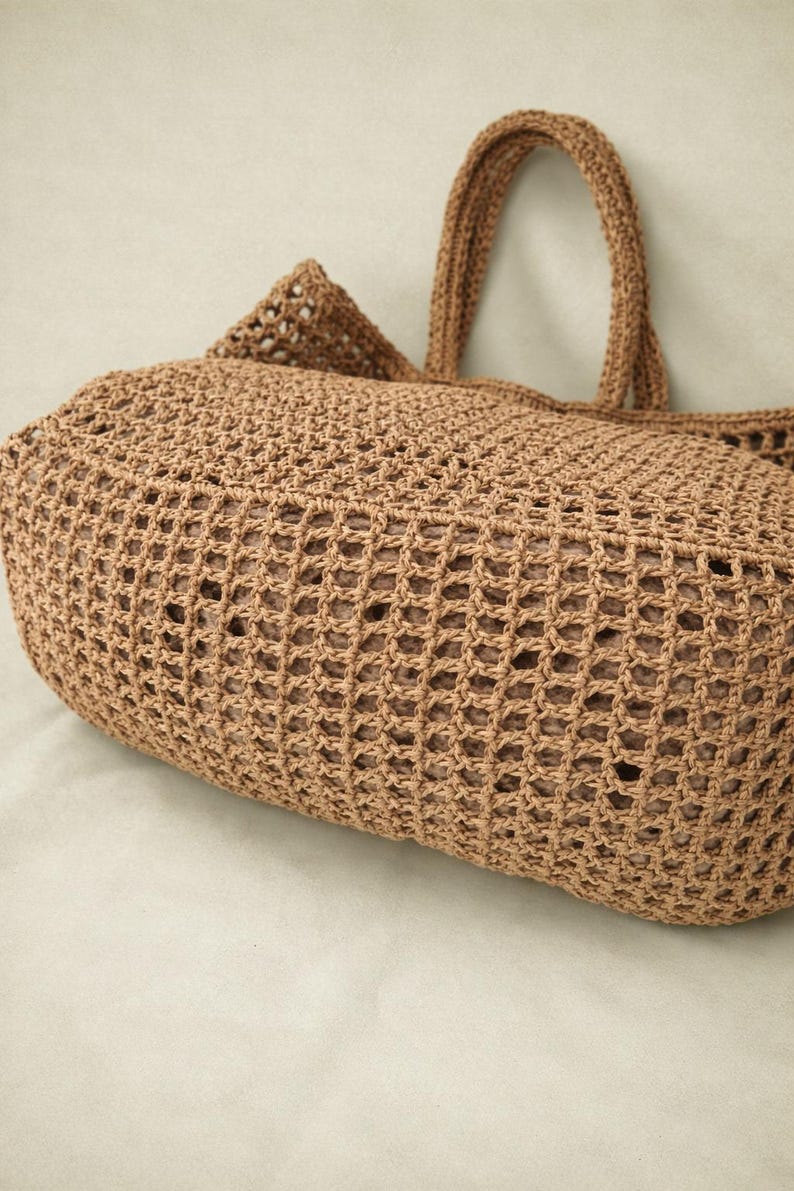 May include: A light brown crochet tote bag featuring an open grid pattern and two sturdy handles. The bag has a rounded, structured base and a visible inner lining that provides a solid appearance through the woven exterior mesh construction.