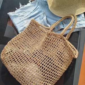 May include: A tan crocheted tote bag with an open mesh pattern and two handles, accompanied by a pair of light blue denim shorts with frayed edges and a tan woven sun hat with a wide brim, all arranged together in a single composition.