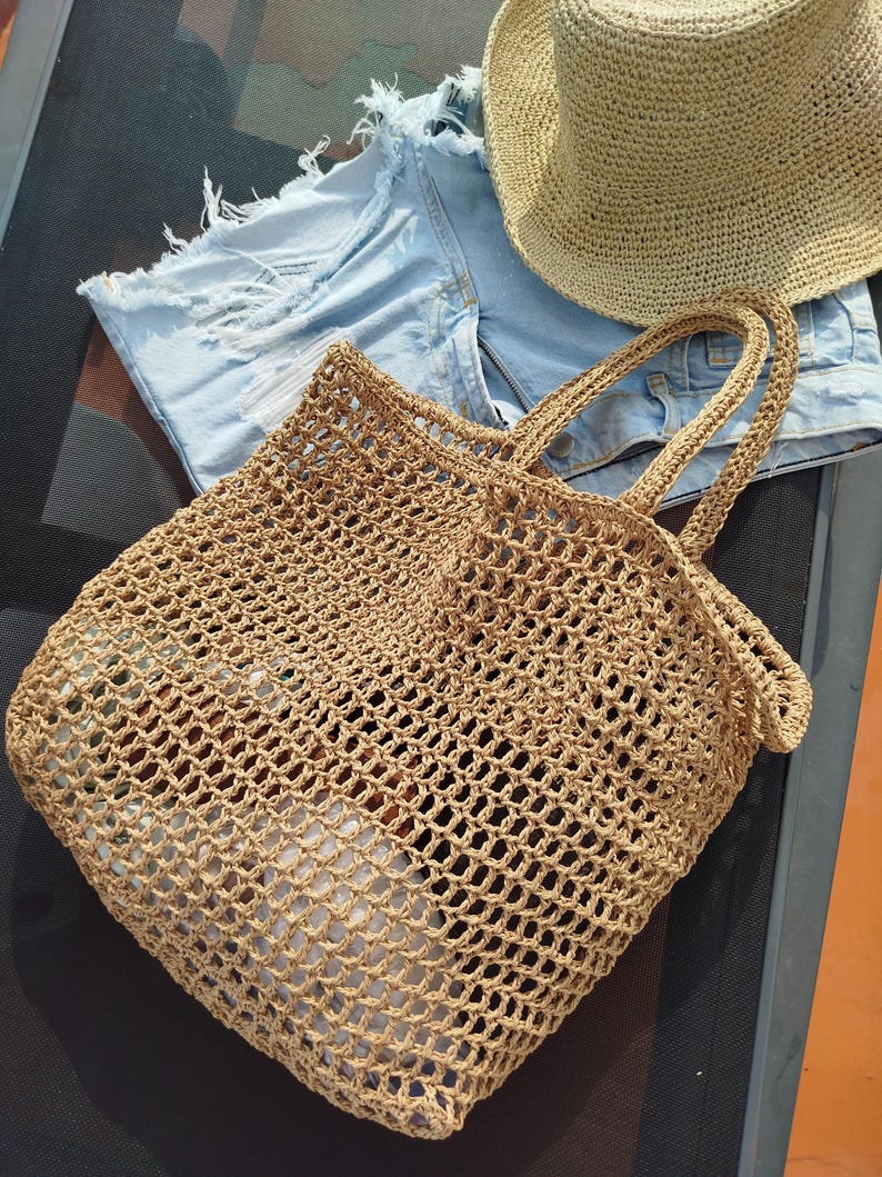 May include: A tan crocheted tote bag with an open mesh pattern and two handles, accompanied by a pair of light blue denim shorts with frayed edges and a tan woven sun hat with a wide brim, all arranged together in a single composition.
