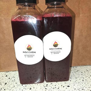 May include: Two clear plastic bottles filled with a dark red liquid, each topped with a black cap. White labels with the text "Deba's Cooking" and a logo are affixed to the bottles. The bottles are on a speckled surface.