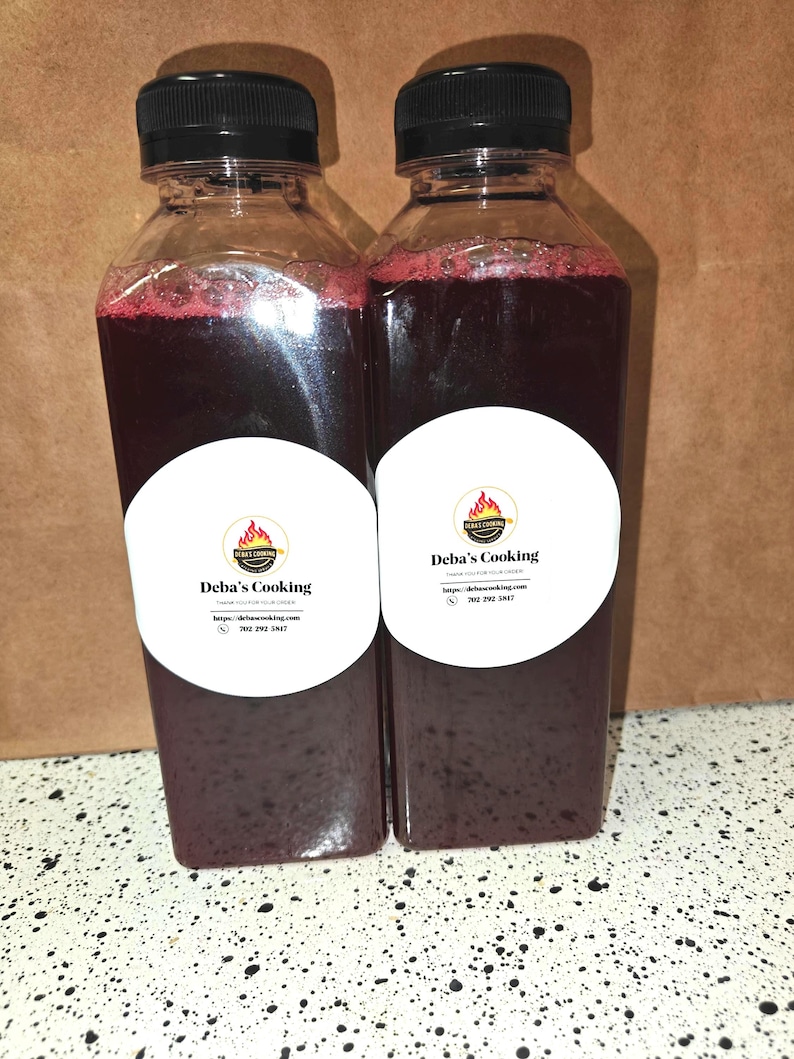 May include: Two clear plastic bottles filled with a dark red liquid, each topped with a black cap. White labels with the text "Deba's Cooking" and a logo are affixed to the bottles. The bottles are on a speckled surface.