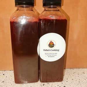 May include: Two clear plastic bottles filled with a dark red liquid, each topped with a black cap. One bottle has a white label with the text "Deba's Cooking" and a logo. The bottles are on a speckled white surface.