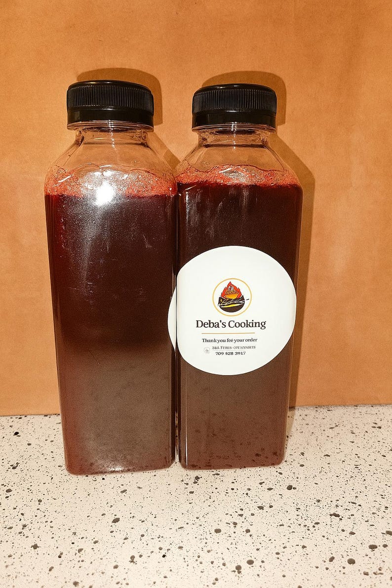May include: Two clear plastic bottles filled with a dark red liquid, each topped with a black cap. One bottle has a white label with the text "Deba's Cooking" and a logo. The bottles are on a speckled white surface.
