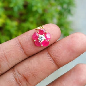 May include: A flower-shaped pendant with a gold bail. The pendant features a central, round, clear gemstone surrounded by six vibrant, oval-cut, red gemstones. The pendant is held between fingers against a blurred green background.