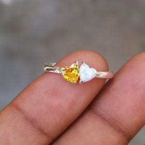 Puede incluir: Un anillo de plata con dos piedras preciosas en forma de corazón. Una es amarilla, la otra es un ópalo blanco. El anillo se sostiene entre los dedos, destacando el diseño y el contraste de color. Las piedras están engastadas en una banda de plata.