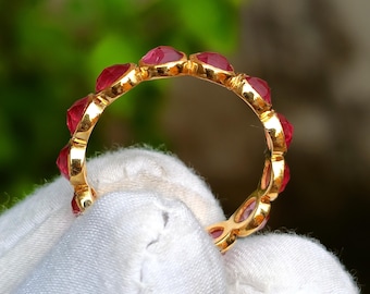 Könnte beinhalten: Ein goldener Ring mit einer Reihe leuchtend roter Edelsteine. Der Ring wird vor einem weißen Stoffhintergrund mit einem verschwommenen grünen Hintergrund präsentiert. Die Edelsteine sind gleichmäßig um das Ringband angeordnet.