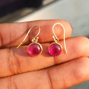 Puede incluir: Un par de pendientes colgantes dorados con piedras preciosas redondas de color rosa brillante. Los pendientes tienen un diseño sencillo con cierre de gancho. Las piedras están engastadas en un bisel dorado.