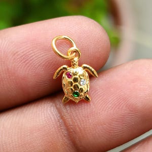 Puede incluir: Un dije de tortuga dorada con un lazo para un collar o pulsera. El caparazón de la tortuga está adornado con piedras preciosas rojas, blancas y verdes. El dije se sostiene entre los dedos.