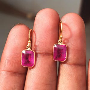 May include: A pair of gold-colored drop earrings, each featuring a rectangular, vibrant pink gemstone. The gemstones are set in a gold bezel, and the earrings have a simple, elegant design. The earrings are shown against a blurred background.
