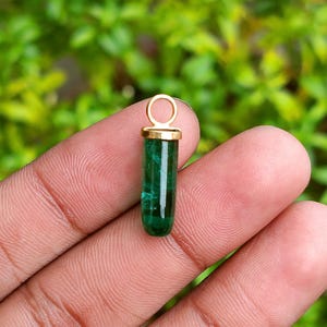 Op de afbeelding: Een kleine, cilindervormige hanger met een levendige groene edelsteen. De edelsteen is gevat in een goudkleurige metalen zetting met een lus aan de bovenkant voor een ketting. De hanger wordt in de hand van een persoon gehouden tegen een vage groene achtergrond.