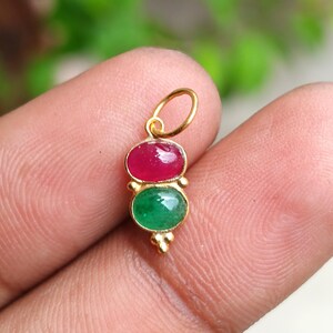May include: A gold pendant featuring two oval gemstones: a ruby on top and an emerald below. The pendant has a small loop for a chain and decorative gold accents. The gemstones are set in a gold bezel.
