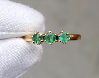 May include: A gold ring featuring three round, vibrant green emeralds. The gemstones are set in a row, creating a classic and elegant design. The ring's band is a warm gold color.